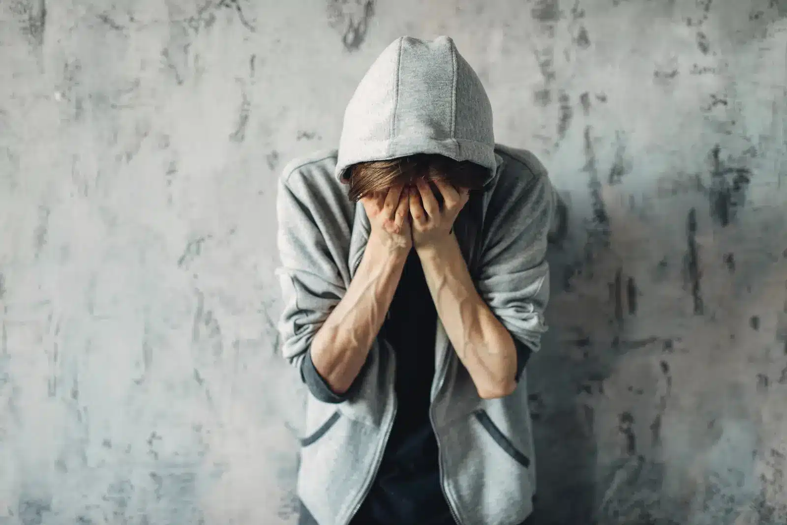 Young man struggling with addiction, wearing a gray hoodie and covering his face with his hands in distress.